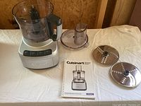 Full view of assembled processor base with bowl and lid, two steel discs, and user manual laid out on table.