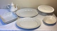 Collection of white serving dishes including a teapot, oval platters, square dish, small bowl, and a milk glass cake stand, displayed on a table.