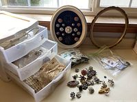 Overall collection showing clear plastic storage drawers with lace trims, a round framed wall art decorated with buttons, an empty wooden oval frame, and scattered brooches on a table.