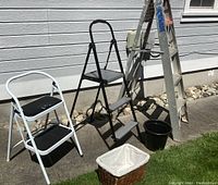 Image showing all items in the lot including two step ladders, 5 ft aluminum ladder, cleaning bucket, and basket arranged outside against a house wall.