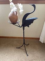 Front angle of metal heron plant stand holding a terra-cotta potted plant with white flowers.