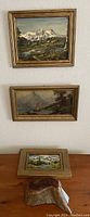 Two framed paintings on wall above table holding third painting and wood box