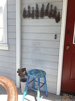 View of the salmon and evergreen trees metal wall art mounted on exterior wall above blue stool with rusty flower decor in bucket