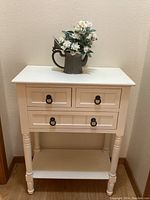 Front view of the white painted wood nightstand with three drawers and black ring pulls, along with a vintage metal pitcher on top containing artificial white roses.