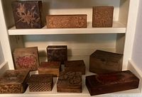 Photo showing 12 pyrography floral wooden boxes of various sizes and shapes on shelves, featuring natural wood tones and some painted accents.