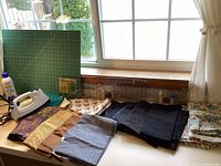 Assorted quilting fabrics folded on a table next to a household iron and a cutting mat near a window.