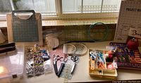 Wide view of sewing and quilting tools laid on table showing Gingher scissors, pins in plastic box, rulers, project board and cutting mat.