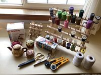 Wide view of sewing notions on a table, including thread racks, scissors, rotary cutters, pin cushions, thread spools, and pin cushion box.