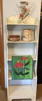 Full view of white painted wooden shabby chic shelf with three shelves. On the top shelf is a metal floral candlestick holder, middle shelf holds two bark baskets, bottom shelf displays quilted floral wall hanging.