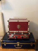 Three stacked vintage cases: a large navy blue metal suitcase base with brass accents, a medium red checkered metal suitcase with chrome details in the middle, and a small red box with white trim on top.
