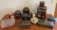Wide view of all 11 vintage decorative metal tins arranged on a table with various floral and oriental designs.