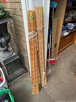 Photo showing two rolled up wicker blinds leaning against a wall in a garage or storage area, tied with pull cords for rolling.