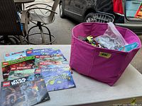 Photo of pink fabric bin filled with assorted loose LEGO bricks and small parts alongside multiple LEGO set instruction booklets, on an outdoor table near chairs and a car.