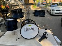 Photo of First Act Discovery kids drum set showing the front with bass drum, two tom-toms, hi-hat cymbal, and pedal on an outdoor table.