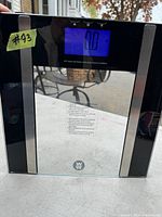 Photo of a black and transparent digital bathroom scale with blue lighted display showing '0.0'. The scale features silver vertical strips on the sides and a Weight Watchers logo in the center front. The display indicates it can measure weight in both lbs and kg.
