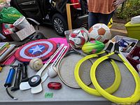 Photo showing a variety of sports equipment on a table including soccer balls, baseballs, badminton rackets, tennis racquet, hockey stick blade with puck, pump, and small accessories