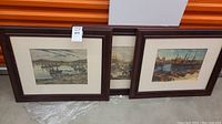 Three framed art prints depicting harbor scenes with boats, placed side by side on a concrete floor.
