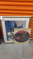 Two framed art prints leaning against an orange storage door on concrete floor; one floral, one satellite image.