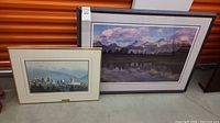 Two framed art prints leaning against an orange storage unit. Larger print shows mountain range with water reflection, smaller print shows city skyline.