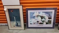 Two framed floral prints side by side against an orange storage door background. One is a single red flower in a white vase, the other is a white cyclamen flower print.
