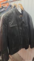 Black leather jacket hanging on a rack, showing front side with quilted shoulder details and zippered pockets.