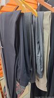 Five pairs of men's dress pants hanging on wooden hangers against an orange background, showing different shades of black, dark grey, medium grey, light grey, and beige.