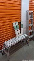 Photo showing the 5-foot aluminum step ladder, folding aluminum bench, and three packages of new window blinds leaning against orange storage unit doors.