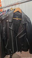 Black leather jacket displayed hanging on rack, showing front view with metal zippers and snap buttons.