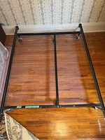Top view of black metal bed frame rail on wooden floor, showing rectangular structure with side and center support bars and four rollers at corners.