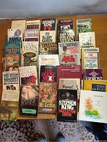 Full view of the assortment of softcover books arranged on a wooden table, shows various titles by Stephen King, Harold Robbins, John Steinbeck and others.