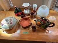 Photo showing variety of ceramic serving dishes, teapot, cups, and glass lamp shades all arranged on a wooden surface under daylight.