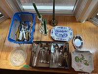 Overview of cutlery pieces, serving dishes, and candlesticks arranged on floor near window.