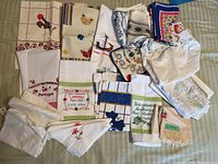 Full assortment of kitchen linens including tea towels, apron, pot holders spread out on a bed showing various patterns and embroidery styles.