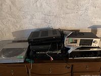 Overview of stereo equipment including turntable, VCR, DVD player, stereo receiver, and cassette player on wood dresser against white brick wall.