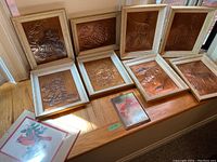 Eight framed embossed copper artworks arranged on a wooden bench, showing diversity of subjects and wood frames with decorative edges.