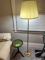 Full view of the floor lamp beside the small side table, showing lamp lit with beige pleated shade and green top of the table