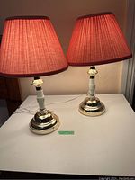 Two matching table lamps with brass bases, white segmented stems, and pink pleated lampshades on a white surface with a beige background.