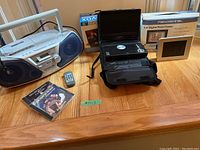 Lot contents showing Panasonic radio/CD player, Audiovox portable DVD player with case, Pandigital digital frame, and remote control on wooden floor.