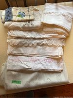 Eight assorted tablecloths stacked. Various crochet lace trims and embroidered details. Mostly white/cream fabrics with delicate patterns.