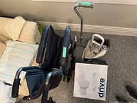 Photo showing folded black travel wheelchair with cushioned armrests and backrest, wide-based metal cane with green rubber handle, and the raised toilet seat box stacked together on carpeted floor