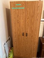 Closed wood grain finish wardrobe showing double doors with dark handles, placed against a wall.