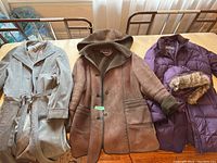 Three women's coats displayed on a table: ultrasuede-like light brown coat, sherpa-style brown coat, and purple Eddie Bauer down jacket with fur hood.