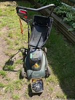 Full view of Yardworks cordless compact lawnmower with attached black grass bag on grassy yard surface, handlebar with red safety cord visible, four plastic wheels with spoked pattern, and a cardboard box holding charger and cords placed in front.