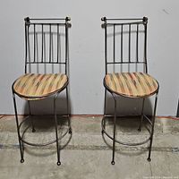 Two metal bar stools side by side showing front view with striped seats and metal backrests with vertical bars and gold-tone finials.