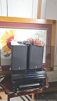 Sony DVD player, two Cambridge Audio speakers, Sony control center stacked and placed on a table with sunflower painting in background. Remote and cables on top.