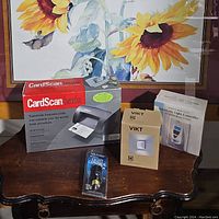 Lot displayed on wooden table with framed sunflower painting background, showing boxed CardScan Executive scanner, IKEA VIKT LED lamp box, Sharper Imager light controller packaging, and automatic light controller manual and device.