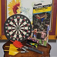 Dartboard with six darts (three metal-tipped and three large plastic tips) placed on a wooden surface, boxed badminton set in background.