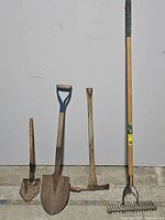 Full set of 4 garden tools including the shovels, pick, and thatch rake upright against a white wall