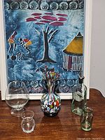 Photo showing all five glass items: brandy snifter, two small clear and green bottles, multicolored decorative vase, and olive oil dispenser bottle with stopper. Blue and red framed artwork background.