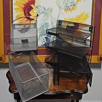 Photo showing all four grey mesh office organizers placed on a wooden table with a floral framed artwork background.